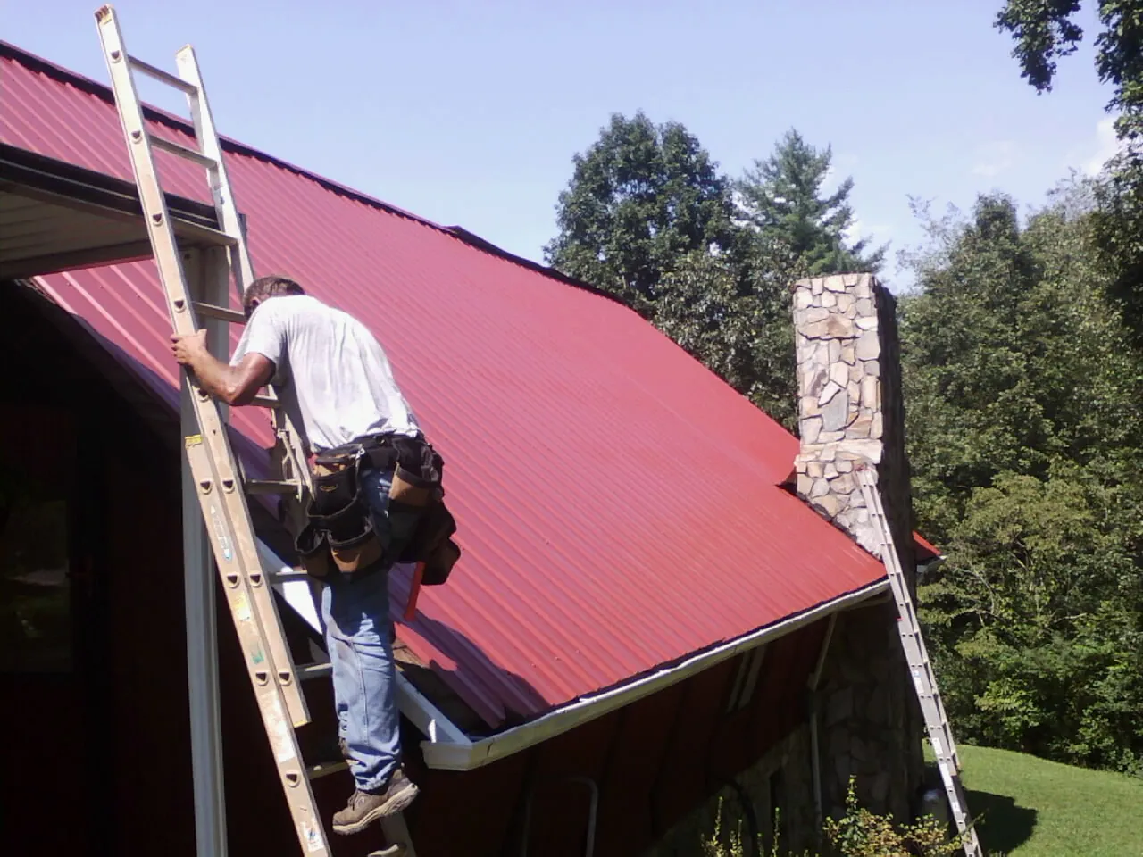 Roofer on a ladder beside a red metal roof and stone chimney — Soffit & Fascia Repair by Roofing Brothers in Philpot